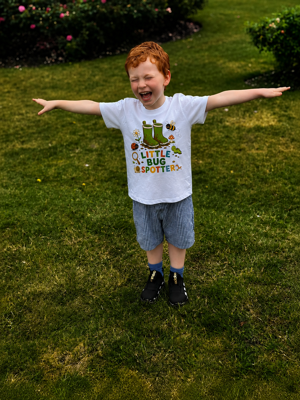 Child wearing a 'Little Bug Spotter' t-shirt standing on grass with arms outstretched