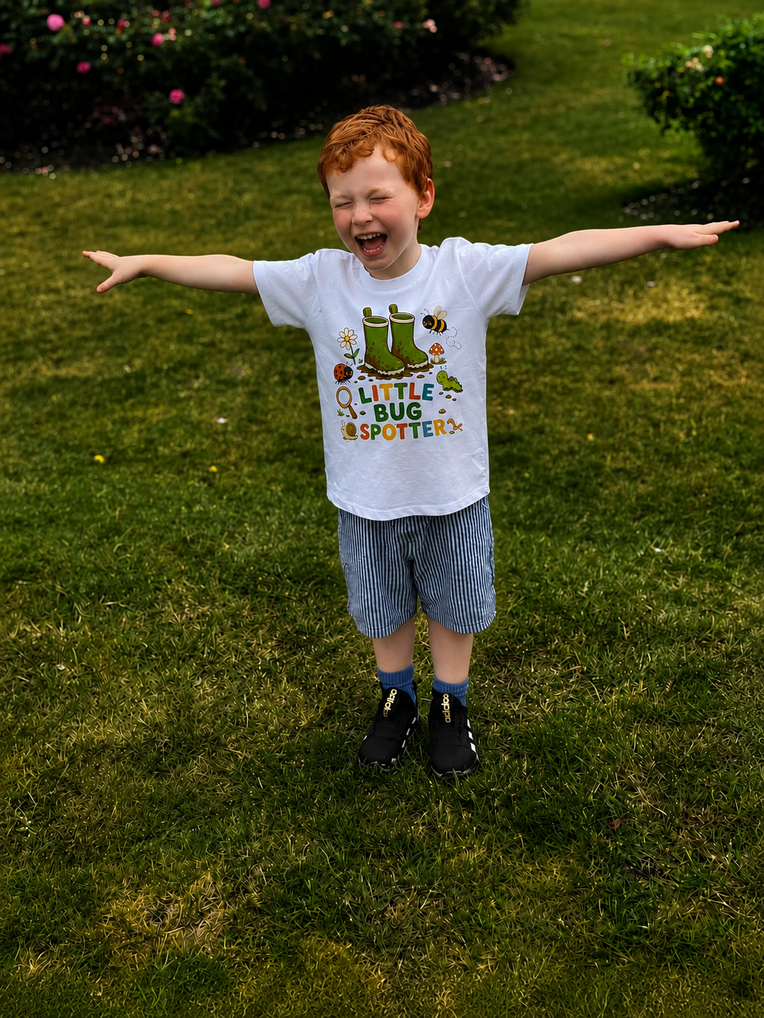 Child wearing a 'Little Bug Spotter' t-shirt standing on grass with arms outstretched