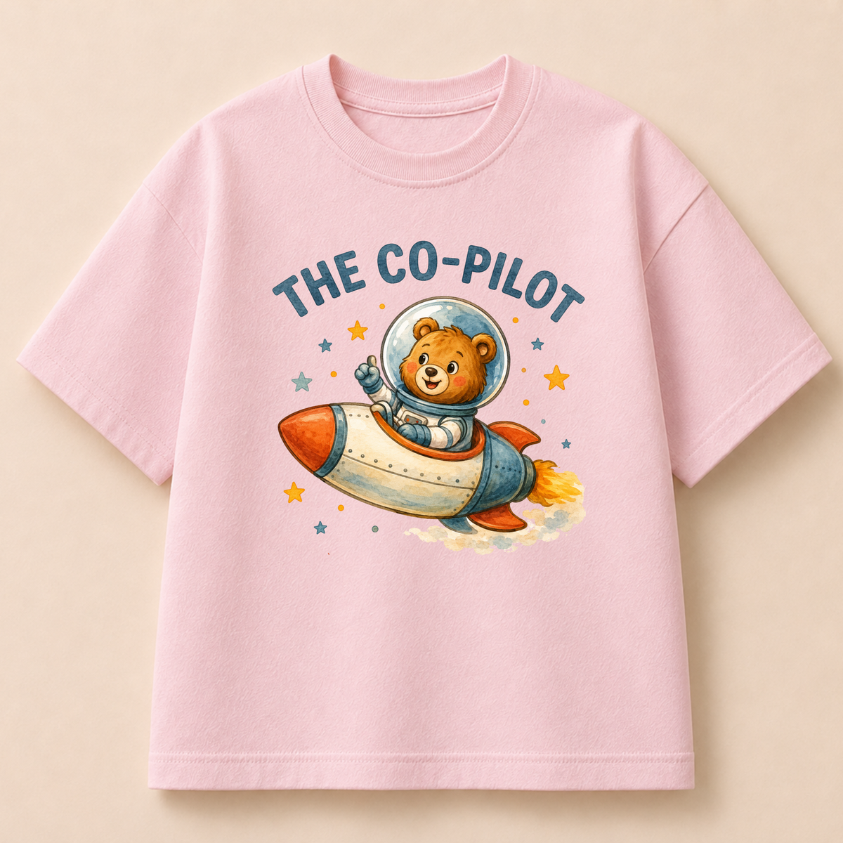 A light pink short-sleeve t-shirt with a cartoon graphic of a teddy bear astronaut flying in a rocket ship, with the text 'THE CO-PILOT' above it, displayed on a solid beige background.
