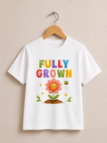 A white short-sleeve t-shirt hanging on a wooden hanger against a beige wall. The shirt features a colorful graphic print with the text 'FULLY GROWN' in rainbow letters, a pink flower with a smiling face, green leaves, yellow stars, and a small bee.