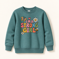 A teal crewneck sweatshirt with a colorful graphic on the front that reads 'I'M A STRONG GIRL' in playful multicolor letters, decorated with cartoon butterflies, flowers, and a bee, shown on a light beige background.