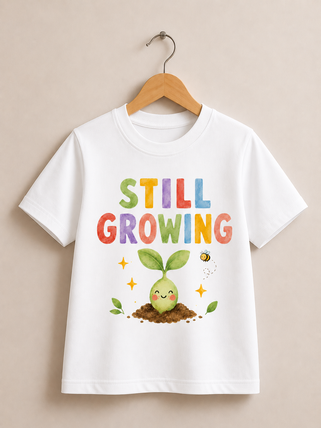 A white short-sleeve t-shirt hanging on a wooden hanger against a beige wall. The shirt features a colorful graphic with the text 'STILL GROWING' in multicolored letters above a cute cartoon green sprout in the soil with a small bee.