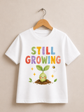 A white short-sleeve t-shirt hanging on a wooden hanger against a beige wall. The shirt features a colorful graphic with the text 'STILL GROWING' in multicolored letters above a cute cartoon green sprout in the soil with a small bee.