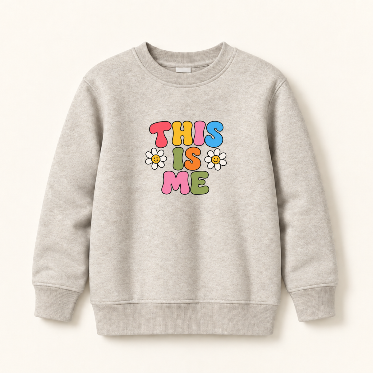 A light heather gray crewneck sweatshirt featuring a retro-style colorful graphic that reads 'THIS IS ME' in bubble letters with small daisy flowers, displayed on a clean off-white background.