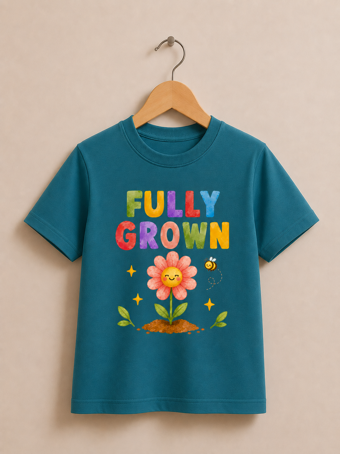 A teal short-sleeve kids' t-shirt hanging on a wooden hanger against a warm beige background. The shirt features a colorful graphic with the text 'FULLY GROWN' in multicolored letters, a smiling pink flower, green leaves, and a small bee.
