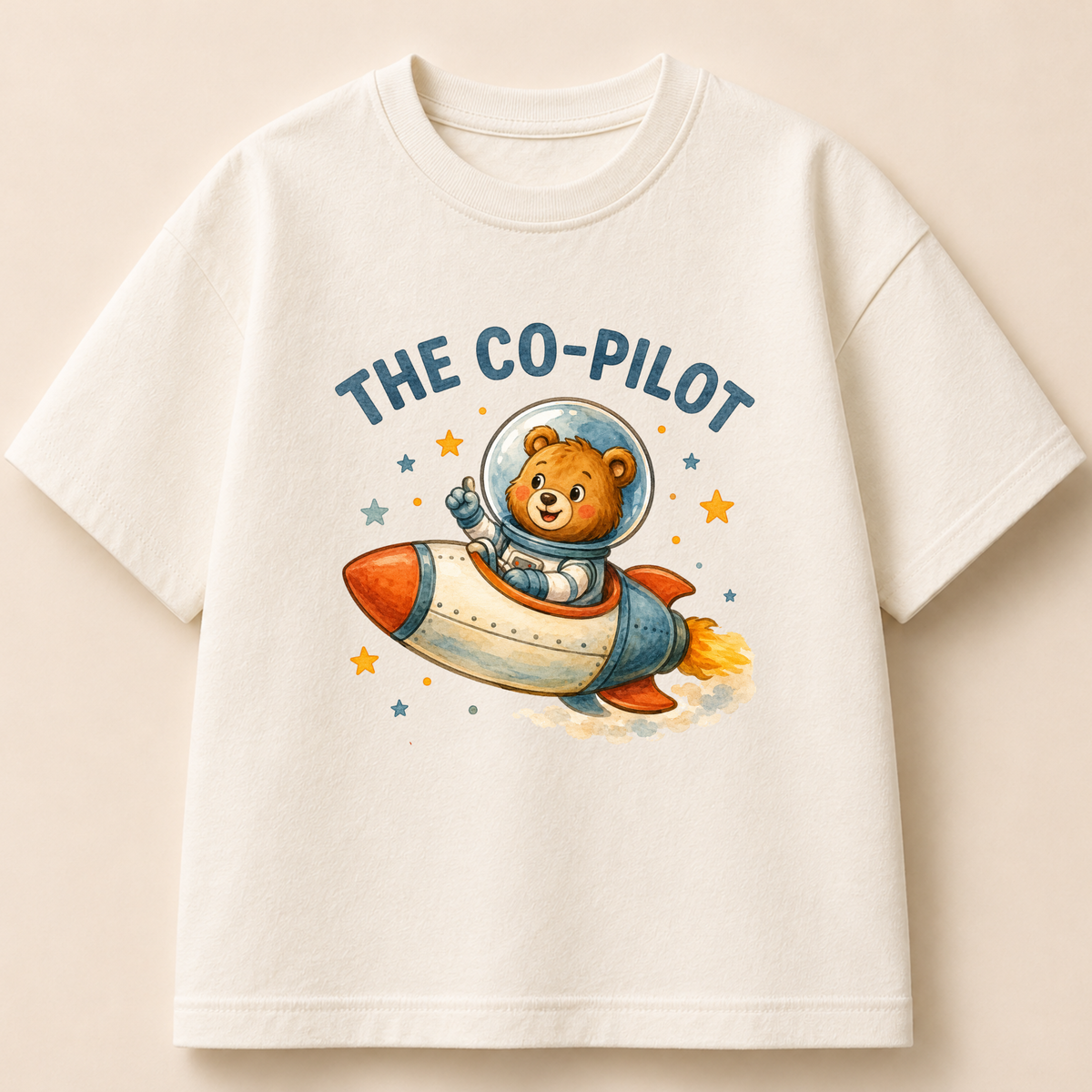 An off-white short-sleeve crewneck t-shirt featuring a cartoon graphic of a teddy bear astronaut flying in a rocket ship with the text 'THE CO-PILOT' on a warm beige background.