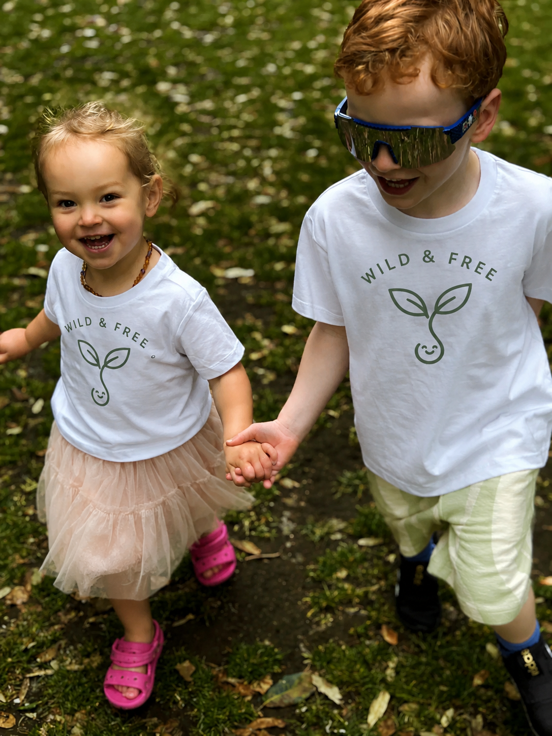 Two siblings wearing 'Wild & Free' t-shirts outdoors on a leafy ground.