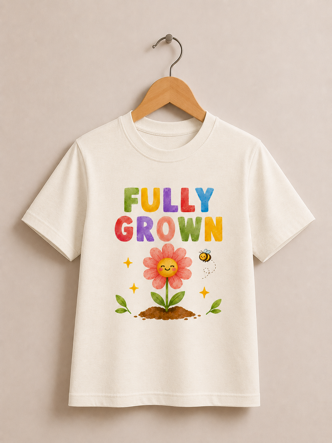 An off-white short-sleeve t-shirt hanging on a wooden hanger against a neutral beige wall. The shirt features a colorful graphic print that reads 'FULLY GROWN' in rainbow letters above a smiling flower, green leaves, and a bee.